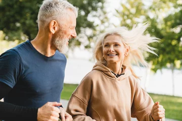 Mature middle aged senior couple running together in the park stadium looking at each other while jogging slimming exercises