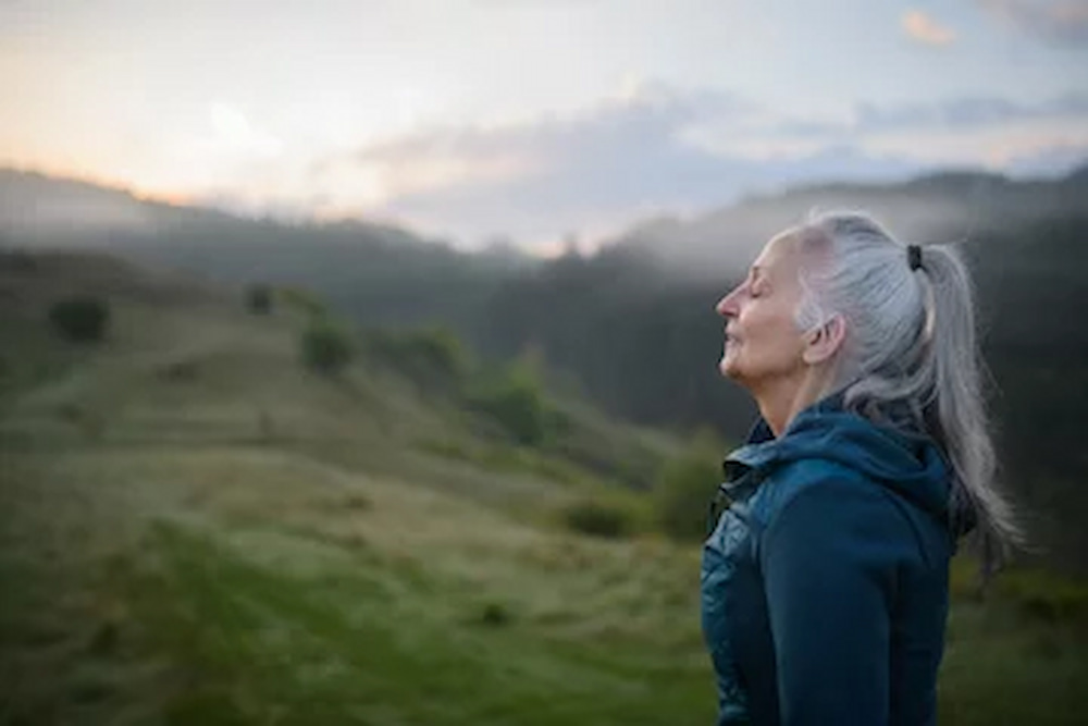 Senior woman doing breathing exercise in nature on early morning with fog and mountains in background
